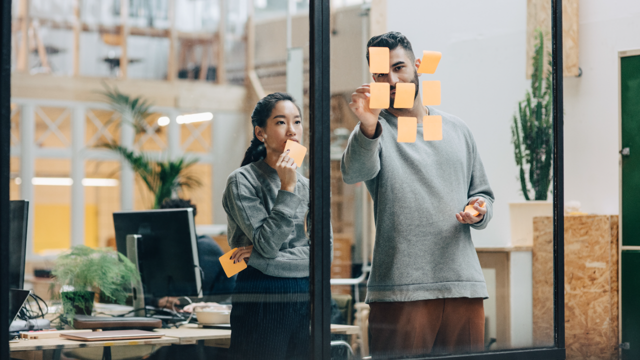 Two colleagues stand in front of a window comparing options on sticky notes.