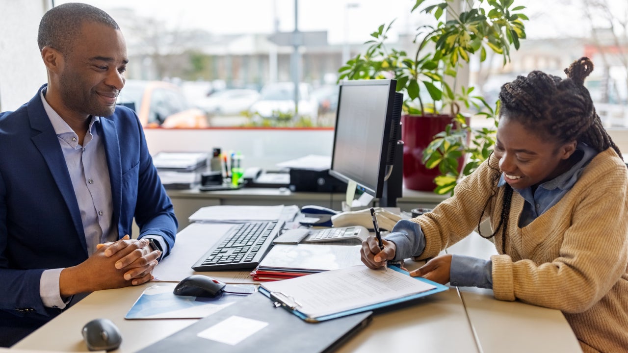 Person filling out forms at a car dealer while a salesperson guides them