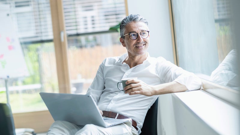 Thoughtful businessman holding coffee cup and laptop looking out through window at office