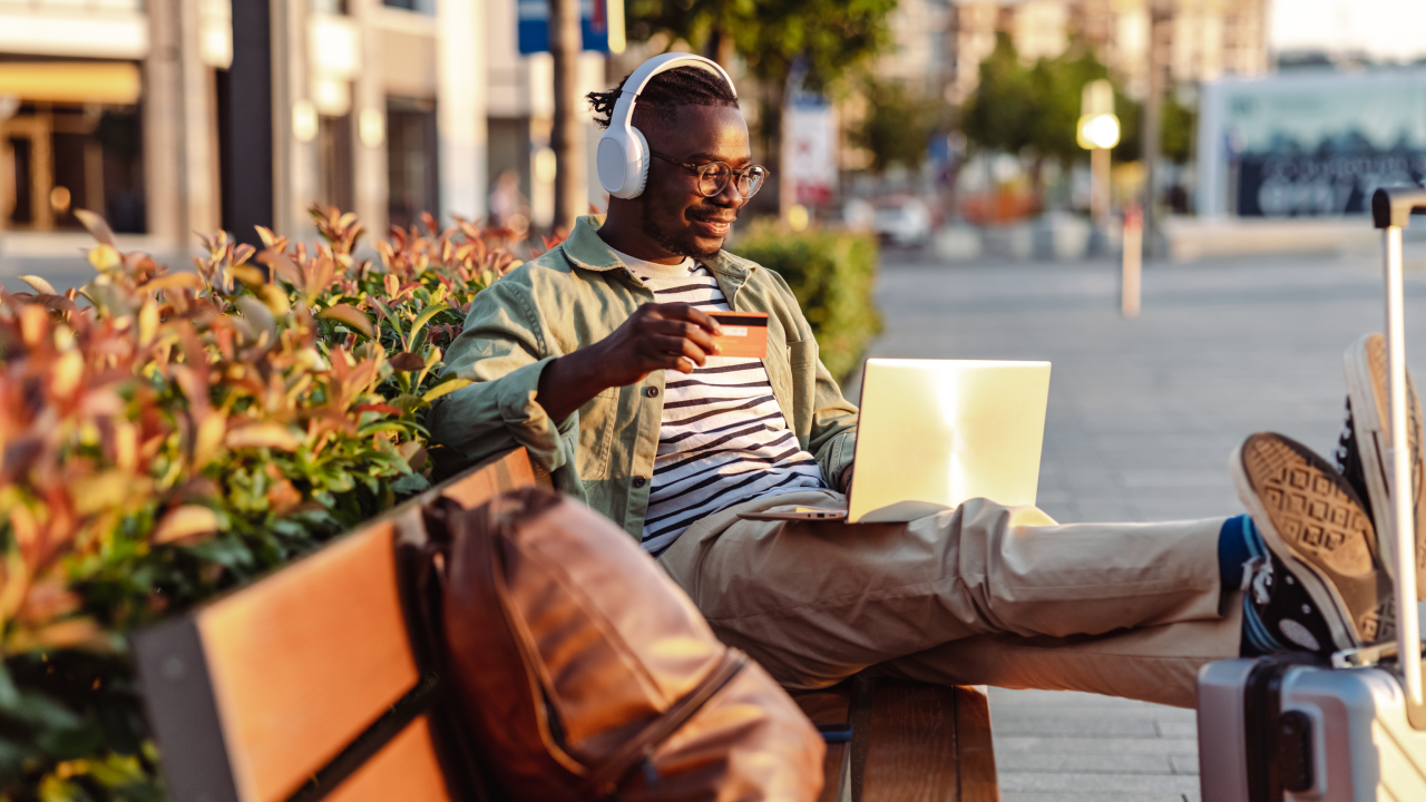 Photo of a smiling African-American man sitting on the bench and holding a credit card. E-commerce activity