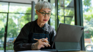 A gray-haired Asian woman, a business owner, sits at a laptop.