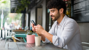 Portrait of a happy Latin American man drinking checking his cell phone at a coffee shop while drinking a cappuccino