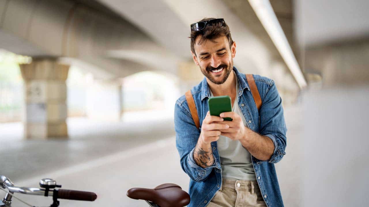 Young man stands next to a bike and smiles at his mobile phone.