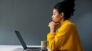 Black woman in an orange shirt viewed in profile looking down at her laptop thoughtfully with her chin on her hands.