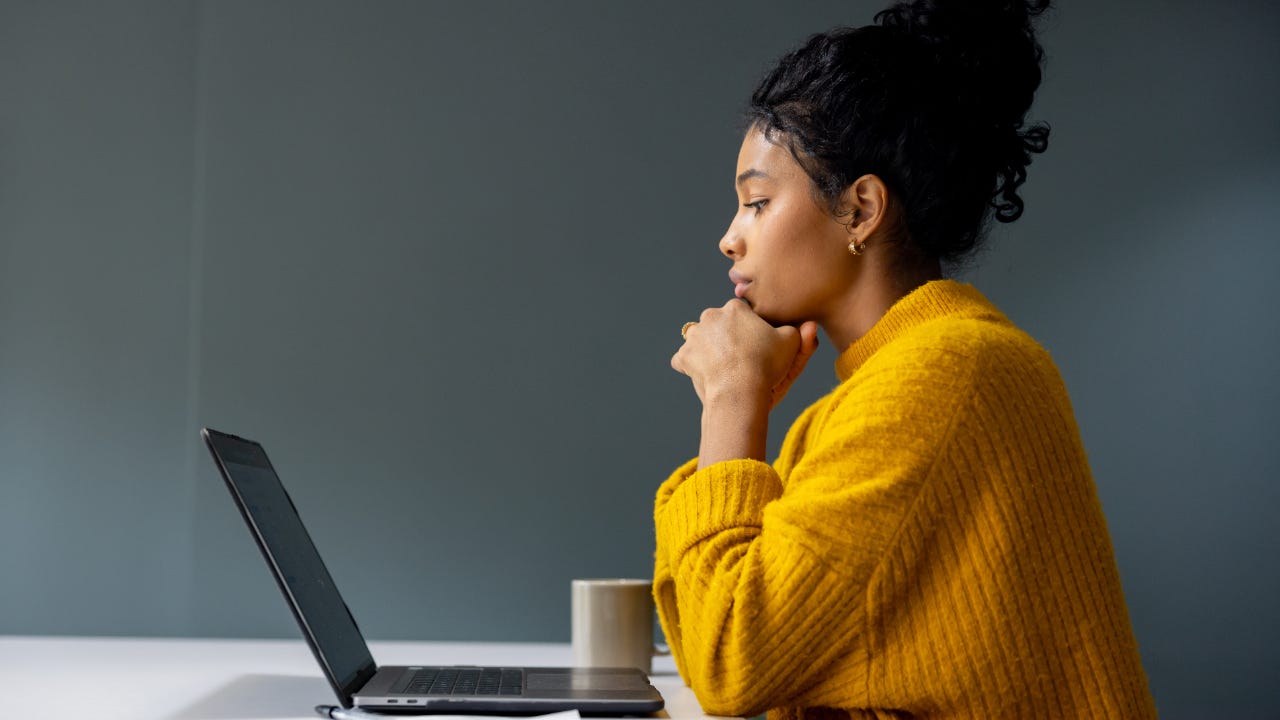 Woman in an orange shirt viewed in profile looking down at her laptop thoughtfully with her chin on her hands