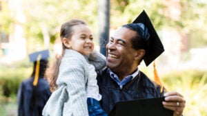 Older graduate smiling at his laughing granddaughter who he is holding