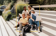 Four college students sitting on outside stairs comparing notes and talking with a yellow illustrated border around them