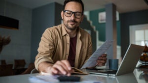 Young man working on finances at home. He has a laptop open and is using a calculator.