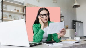 young woman in glasses sitting by her laptop reading a piece of paper in dismay