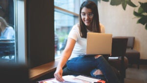 Young woman in glasses works at a low table in a college room. She has a laptop and a cheerful expression.