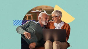 An older couple laughing on a couch, one of them is on a laptop