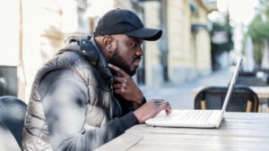 Black man sitting at an outdoor cafe thoughtfully looking at his laptop.