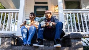 A couple sits on a porch of a house drinking coffee.