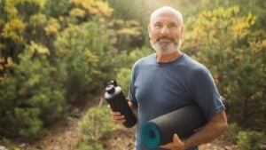 A senior man holds a water bottle and yoga mat.