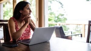 Middle aged woman looks out window sitting at table with laptop in front of her.