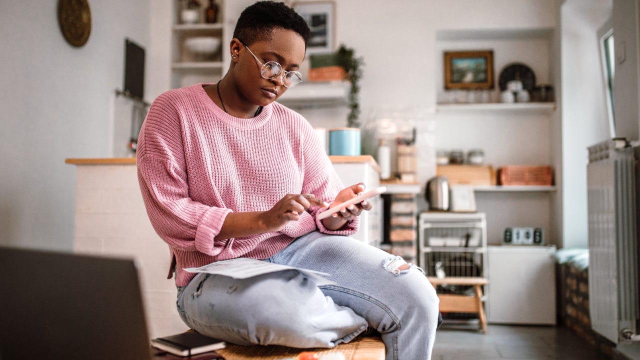 Black woman sitting in her apartment using her phone with her laptop in front of her.