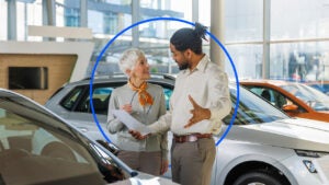 A younger Black salesperson talks with an older white car buyer at a dealership. The image includes a dark blue circle around both individuals.