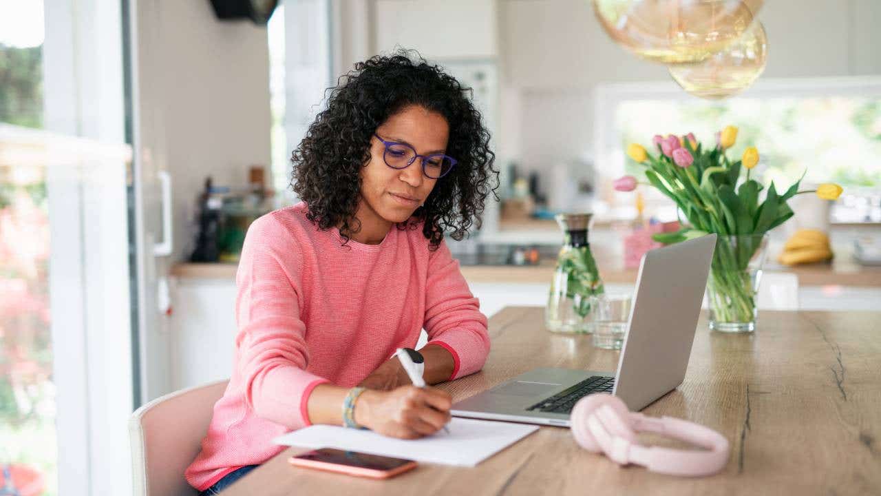 A woman writing notes in her kitchen next to a laptop.