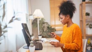 A woman works on her laptop while holding her phone and looking at papers.