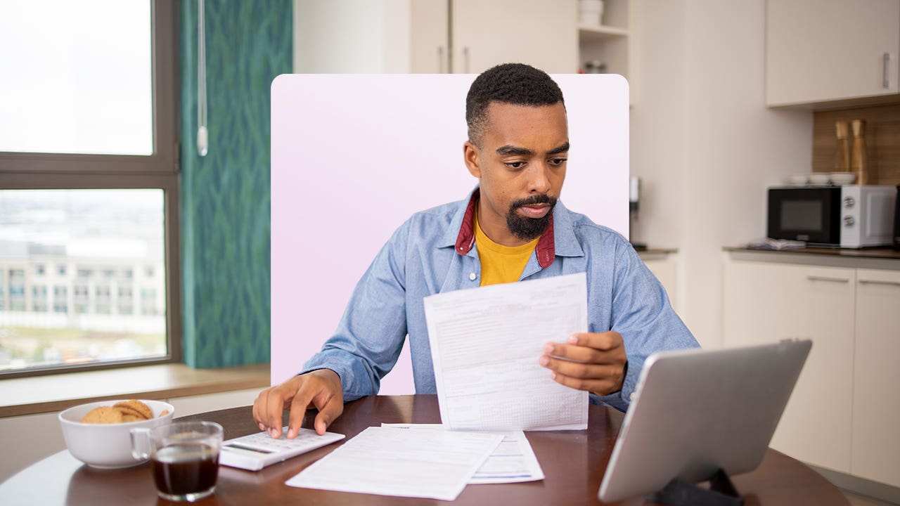 A Black man sits at a table using a tablet and doing calculations.