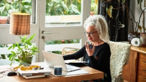 Cheerful senior businesswoman speaking on smart phone in front of laptop