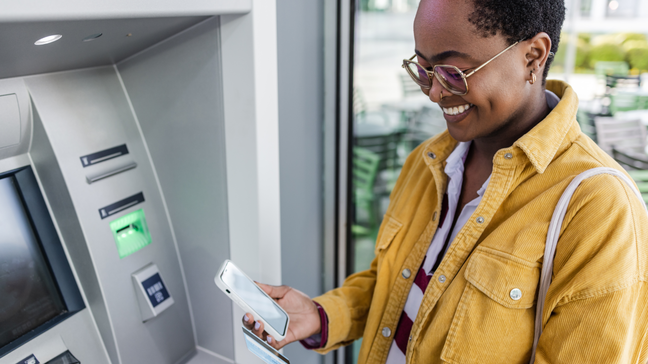 Young woman is holding a credit card and a mobile phone. She is standing in front of the ATM and thinking about the method of withdrawing money.