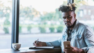 Young Black man sitting at coffee shop and checking his phone while writing on a piece of paper.