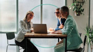 Two women and a man sit at a table and look at a laptop.