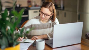 Young white woman working at a laptop and writing in a notebook at her kitchen table.