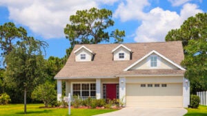 New cozy cottage with garage, driveway, dormer windows, lawn and trees in background.