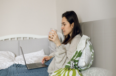 The Caucasian woman is lying on the bed and using a laptop to working online from home while her child is in another room with father