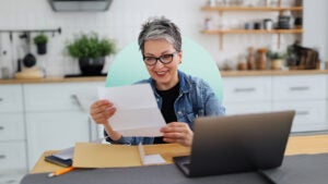 design image of an older woman looking at a piece of paper in front of her laptop