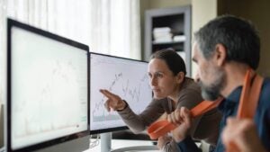 Two people looking are charts on two computer monitors.