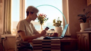 Design element of a person sitting in front of a window using a laptop while holding a credit card in hand