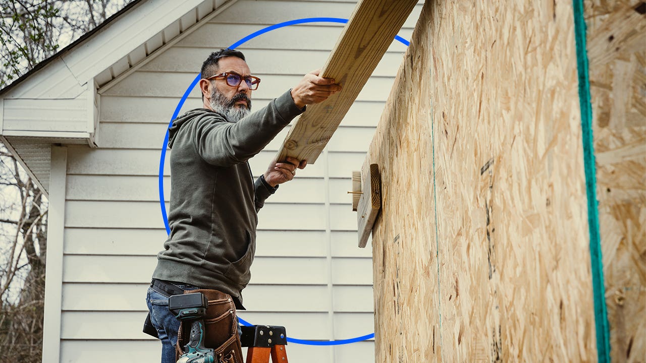 An older white man in glasses putting a board up on a structure with an illustrated blue circle behind him.