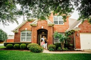 family of 4 posing in front of new red-brick house, green lawn and nice landscaping