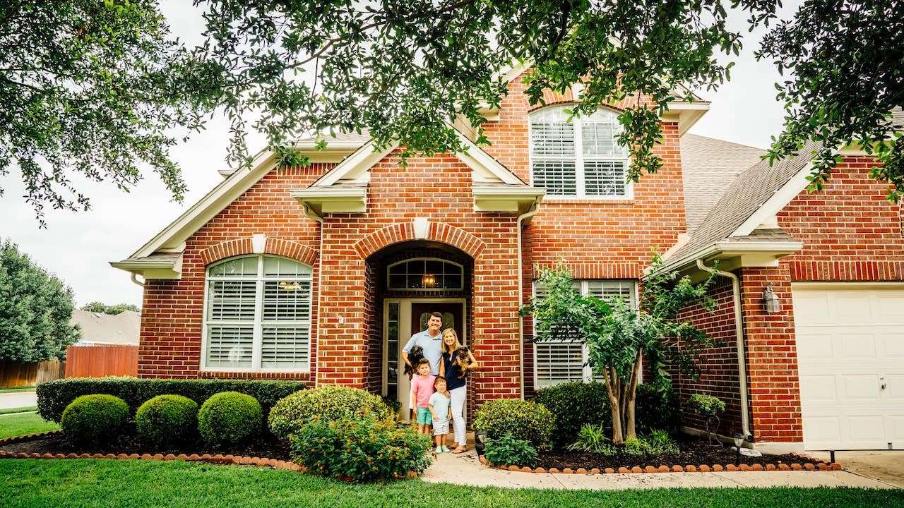 family of 4 posing in front of new red-brick house, green lawn and nice landscaping