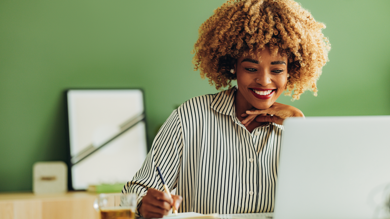 Smiling African American woman reading business report on a laptop and writing notes in notebook while sitting at office desk.