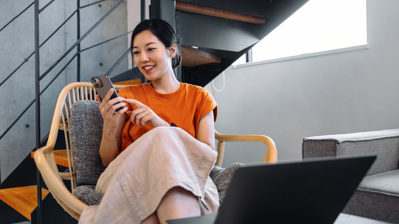 Young Asian woman using smartphone while relaxing on armchair, checking her bank account via online banking and managing her finances. Online personal banking. Wealth management. Digital banking habits. Smart banking with technology