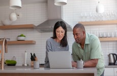 A mixed race couple use a laptop computer to pay bills online, manage budget and prepare tax documents. They are in the kitchen of their home.