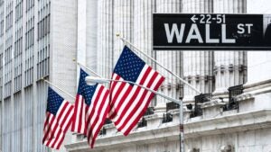 Close-up of the Wall Street street sign. In the background, there are American flags on the exterior of a building with a colonnade.