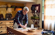 Senior man standing in a kitchen kneading bread dough at a table.