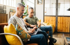 Two gray-haired men sit on a couch using a laptop and going over their home finances in a living room.