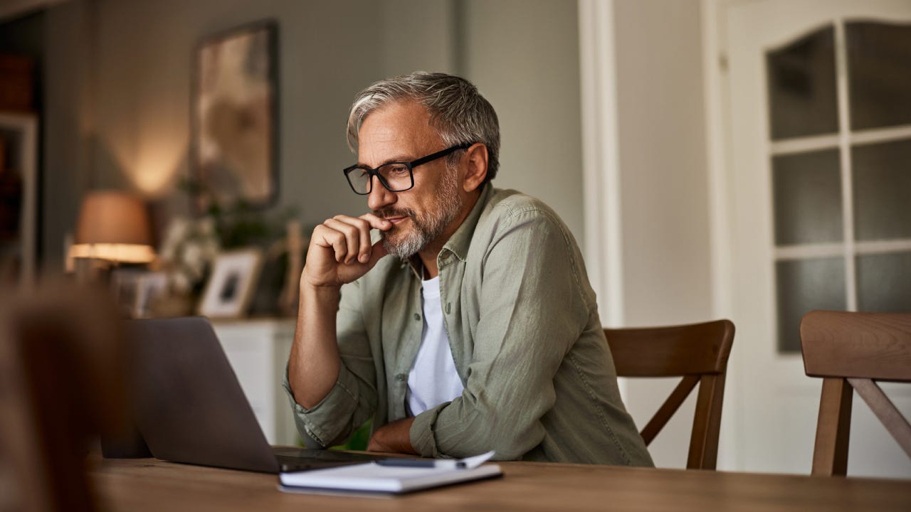 A focused mature male freelancer sitting at the wooden table at home and reading online projects on a laptop.