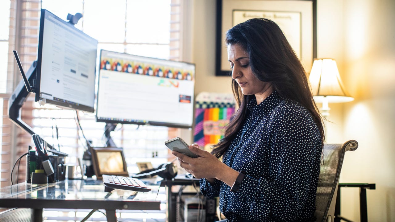 A woman looks at her phone while sitting at her desk with two monitors.