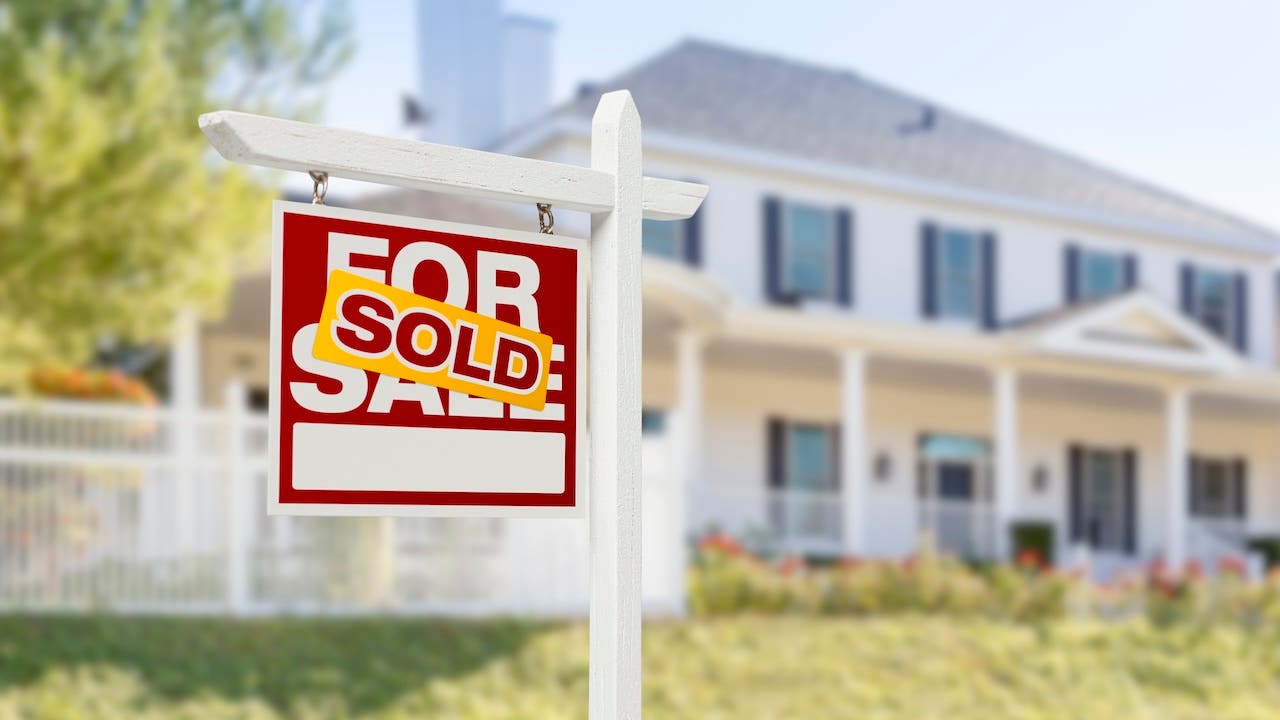 red and yellow "sold" sign in front of white suburban house with black shutters