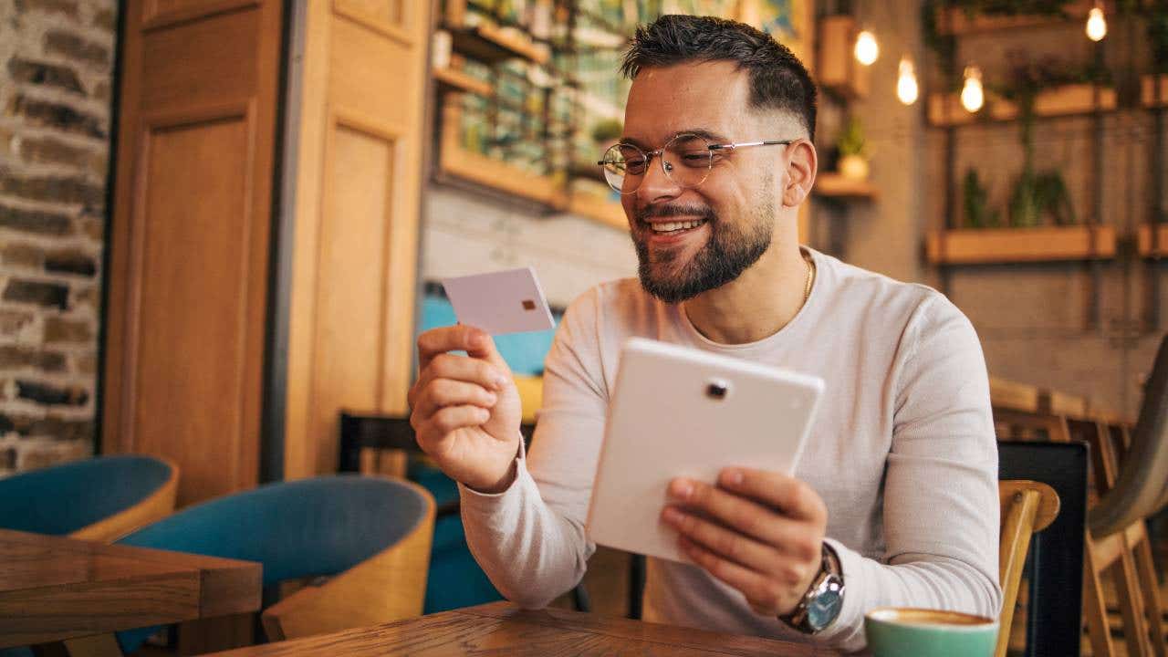 Young man holds a credit card and a tablet sitting in a restaurant.