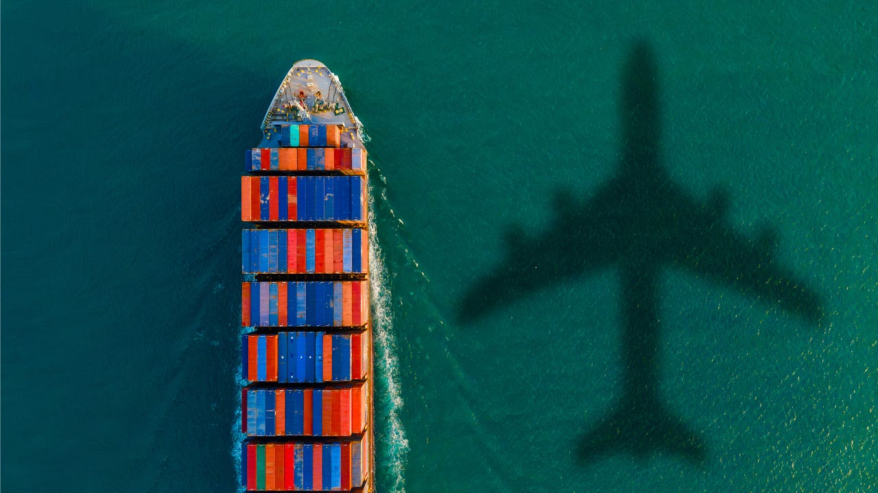 Aerial view of a cargo ship navigating green water with the shadow of a plane overhead