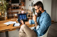 Mid adult man working at home using a cell phone and laptop, while his daughter does her homework in the background of a living room.