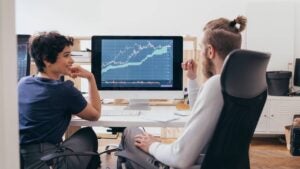 Two people in an office setting sitting next to a computer monitor displaying a stock chart.